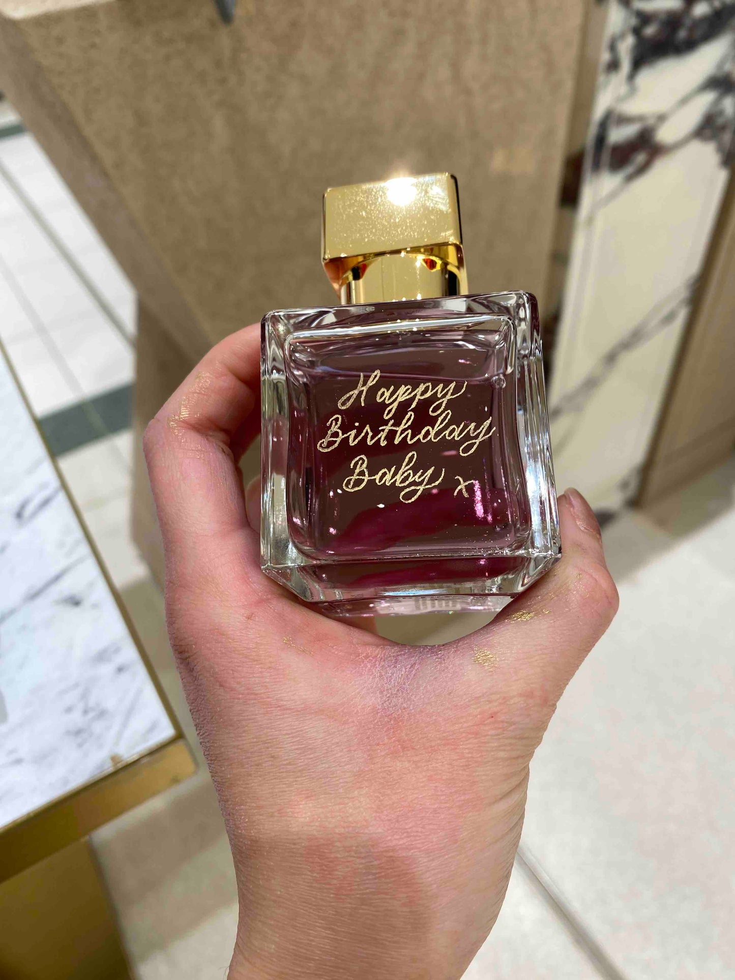 Hand holding a perfume bottle with 'Happy Birthday Baby’ engraved text on a glass surface