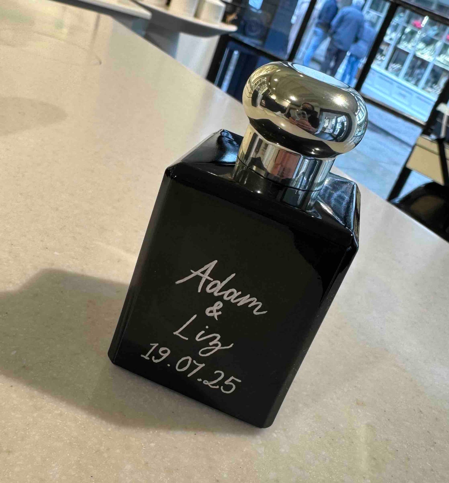 Black perfume bottle with silver cap on a light surface, engraved with 'Adam & Liz 19.07.25'.